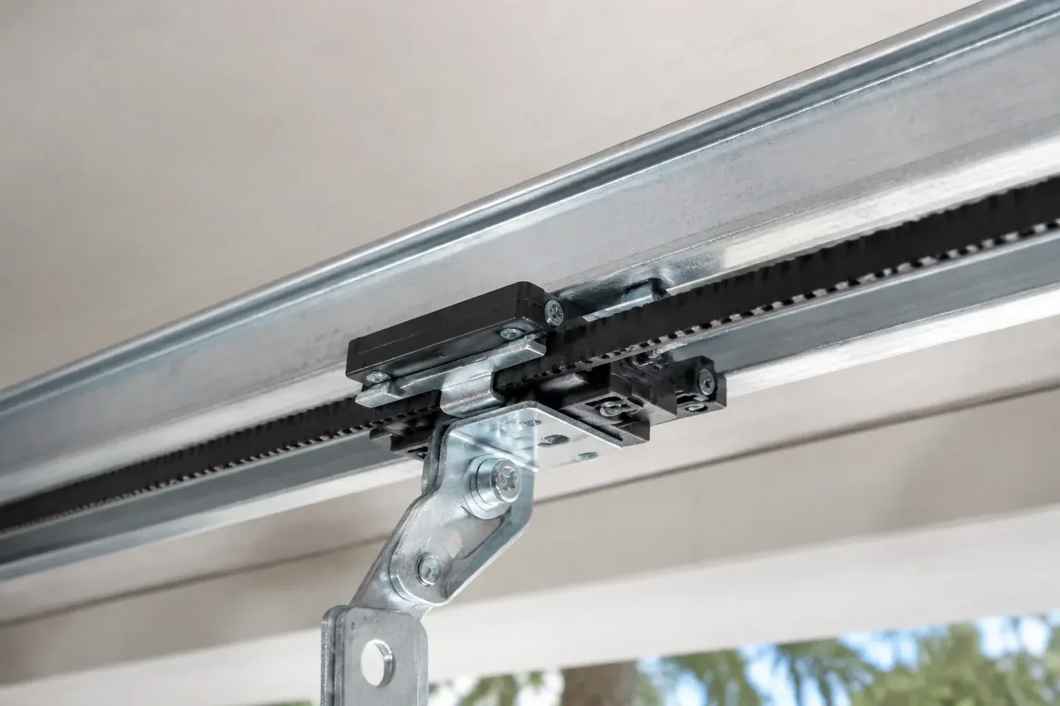 Black plastic trolley and bent galvanized bracket on a steel opener rail with a toothed black belt, against a ceiling. — Belt-Drive Opener Repair, Oshawa