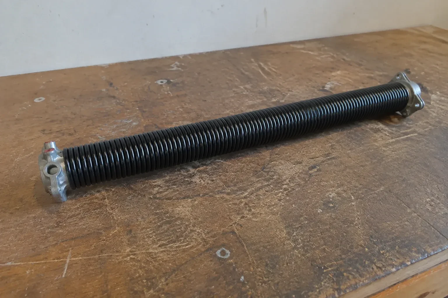 Detached black coil spring with metal end fittings on a workbench. — Torsion Spring Replacement, Oshawa