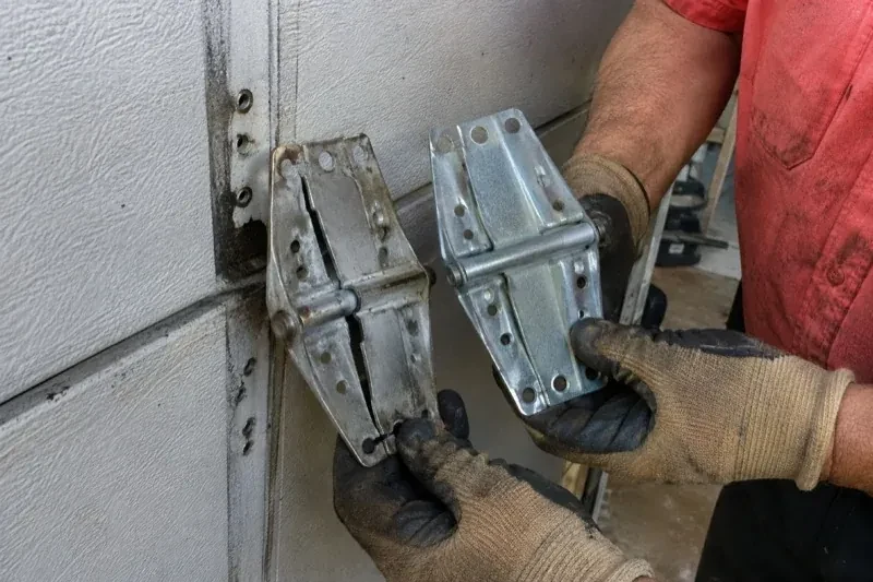Gloved hands holding two metal garage-door hinges side by side, one discolored with a visible crack and the other bright silver. — Garage Door Hinge Replacement, Oshawa