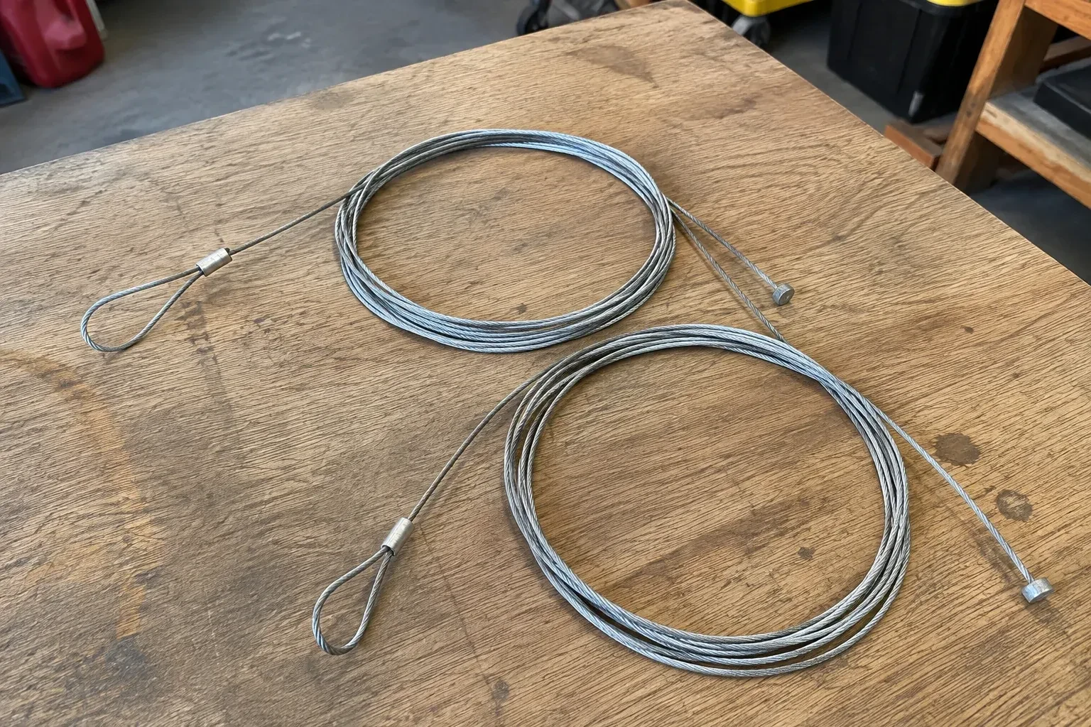 Two coiled steel cables with loop and stop ends on a workbench. — Garage Door Cable Replacement, Oshawa