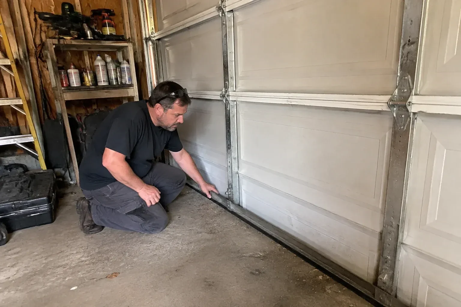 Person kneeling by a white paneled door in a garage, one hand on the black seal along its bottom edge. — Garage Door Roller Replacement, Oshawa