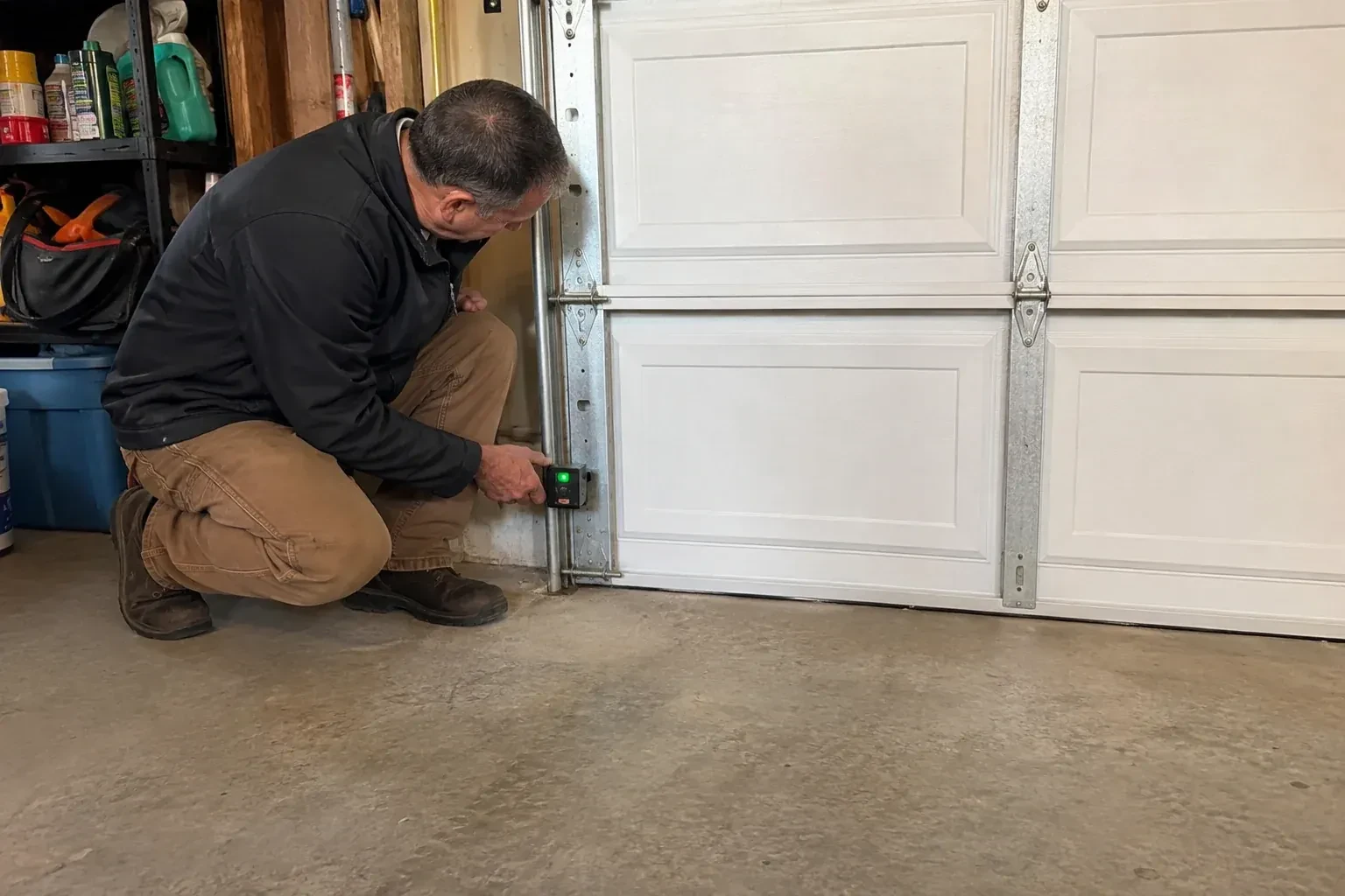 Technician kneeling beside a white paneled garage door, holding a small black box with a green light against the vertical track. — Photo-Eye Sensor Alignment or Replacement, Oshawa